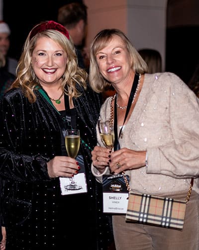 Three people pose and smile at SITE SoCal Holiday Event; a man in a patterned suit stands in the middle with two women in dresses on either side, each holding a drink.