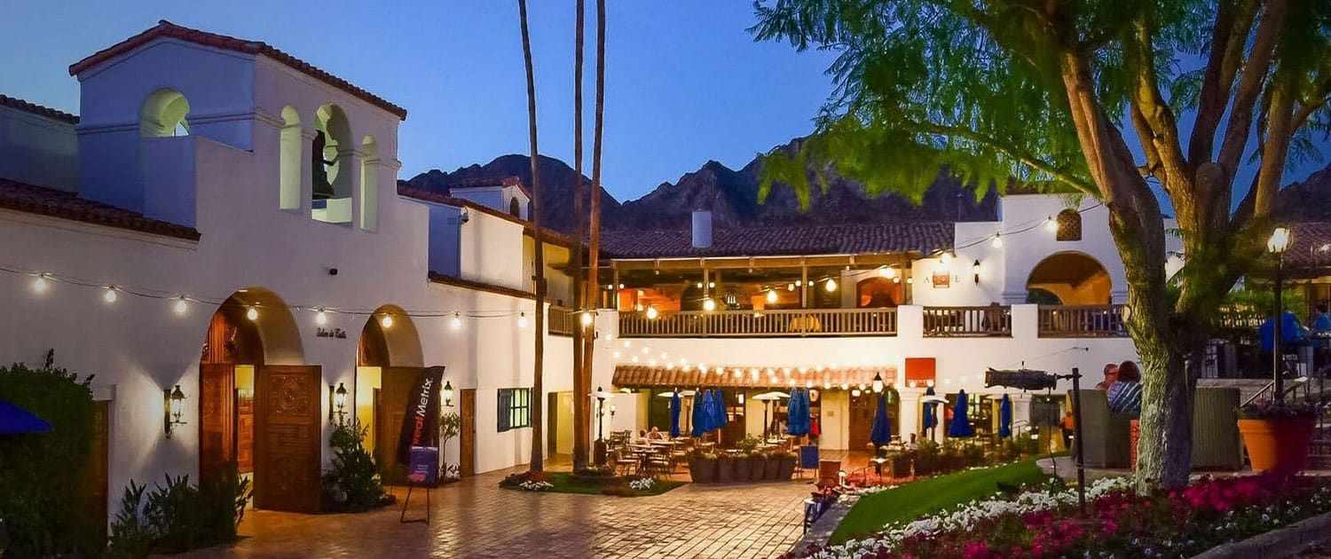 Courtyard of a Spanish-style building at dusk, decorated with string lights, shopfronts, and outdoor seating, with mountains visible in the background.