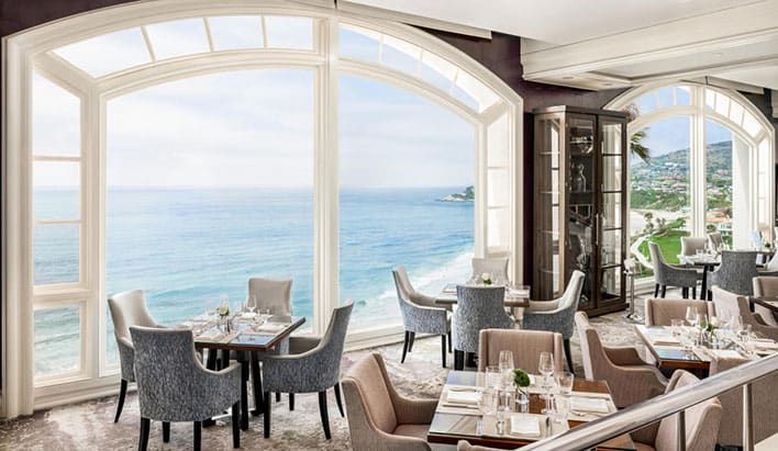 A restaurant dining area with elegant tables and chairs, large arched windows, and a view of the ocean and coastline on a clear day.