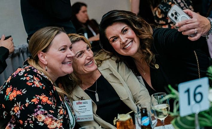 Three women sit together at a table, smiling and taking a selfie during an indoor event. Drinks and a table number 16 sign are visible in the foreground.