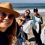 Four people on a beach smile at the camera while holding garbage bags, participating in a Young Leaders Annual Earth Day Beach Cleanup activity .