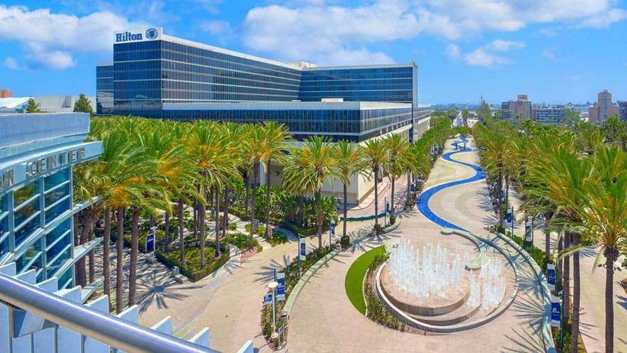 The modern Hilton Anaheim hotel building with glass windows, surrounded by palm trees, wide walkways, and a central fountain under a blue sky.