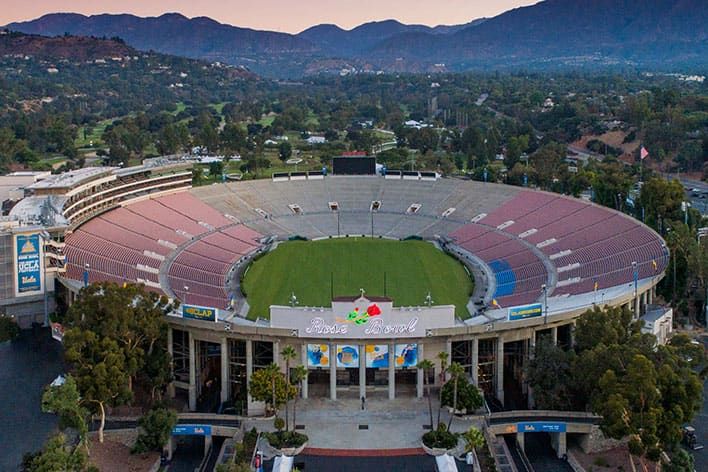 Aerial view of the large, empty outdoor Rose Bowl Stadium with a green field, surrounded by rows of seats, trees, and hills in the background.
