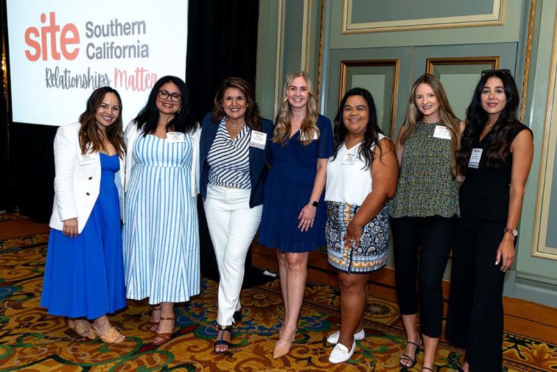 Seven women stand together indoors, smiling at the camera, with a "SITE Southern California" event sign displayed in the background.