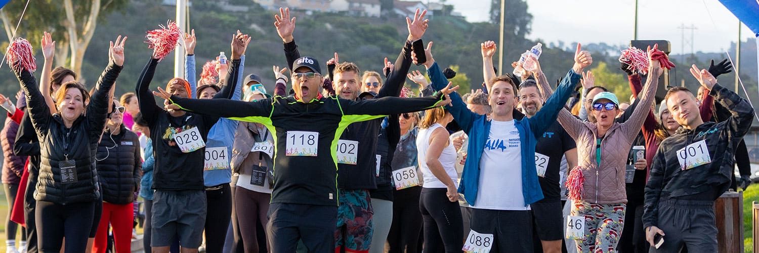 A group of runners wearing race bibs cheer with arms raised at the Holiday Event & Charity Gala 5k, some holding pom-poms.