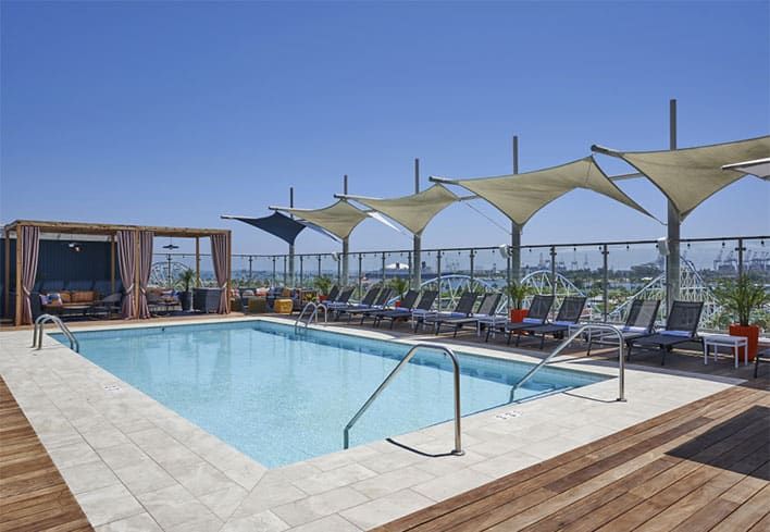 A rooftop swimming pool surrounded by lounge chairs, shaded canopies, cabanas, and glass safety railings, under a clear blue sky.