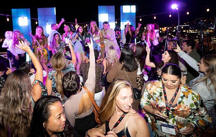 A large group of people gathered at a lively outdoor night event, many holding drinks, dancing, and enjoying music under string lights.