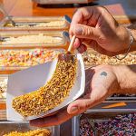 A person holds a chocolate-dipped banana covered in crushed nuts on a stick, with various toppings displayed in trays in the background.
