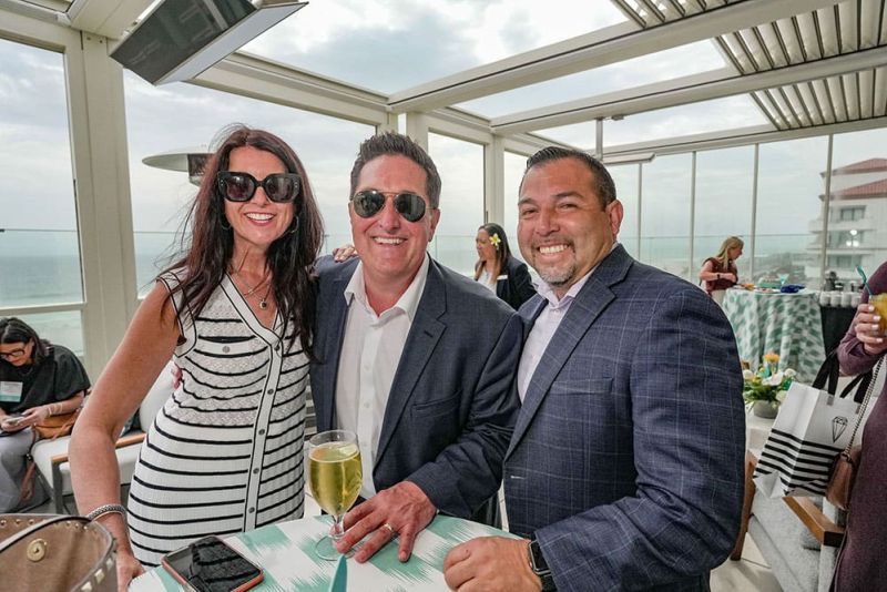 Three people dressed in business attire smile at an outdoor event, standing by a table with drinks, with a view of the ocean in the background.