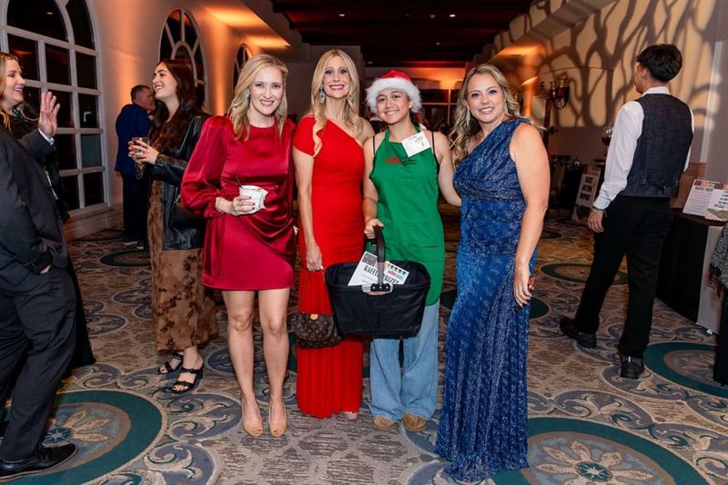 Four women pose together at an indoor event, three in evening dresses and one in a green outfit with a Santa hat holding a basket, with other people mingling in the background.