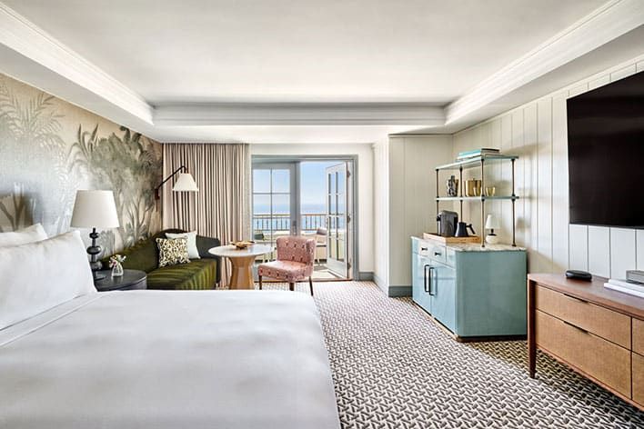 Bright guest room at The Ritz-Carlton, Laguna Beach, with a large king bed, seating area, desk, TV, and a view of the ocean through glass doors leading to a balcony.