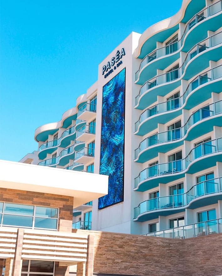 The exterior of Paséa Hotel & Spa, featuring curved balconies and a tall digital art display, under a clear blue sky.