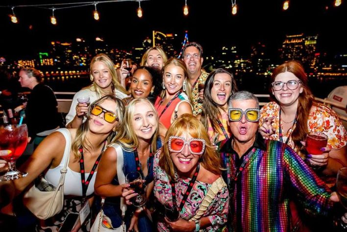 A group of people wearing colorful clothes and glasses pose and smile for a photo at a nighttime outdoor party with city lights in the background.