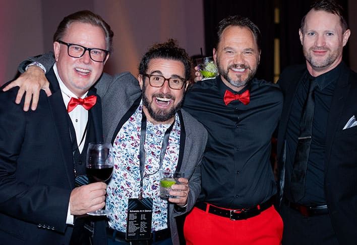 Four men dressed in formal attire, two wearing red bow ties, pose together at an event; one holds a glass of wine, another a cocktail.