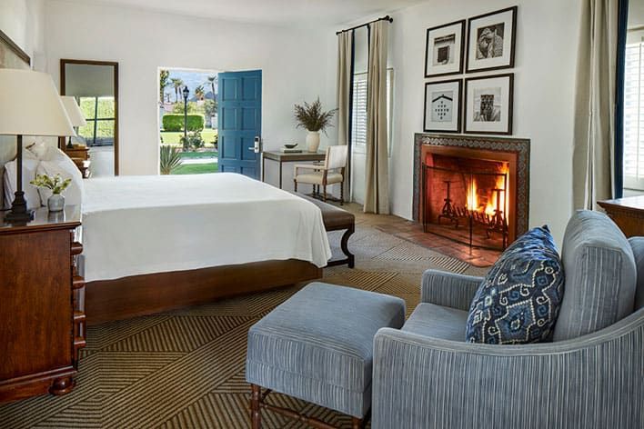 A cozy La Quinta Resort & Club king vintage casita room with a lit fireplace, armchair, wooden bed, desk, and an open door revealing a view of a garden and palm trees outside.