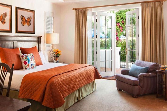 A La Quinta Resort & Spa guest room with an orange bedspread, butterfly artwork above the bed, an armchair, and open French doors leading to a patio with greenery outside.