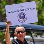 A man wearing sunglasses holds a sign that reads "Welcome SITE SoCal" with a logo beneath the text.