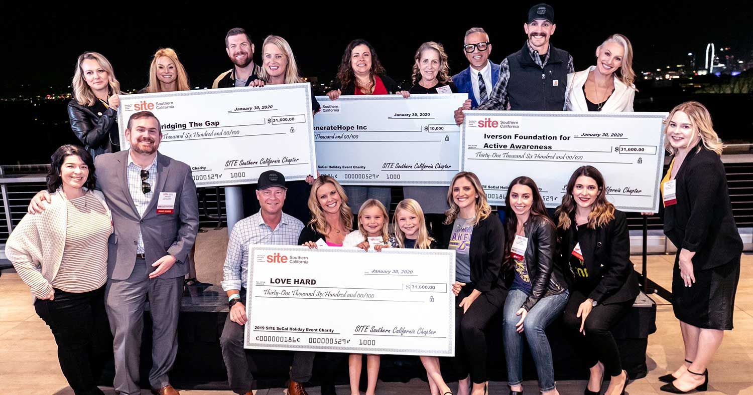 A group of people pose outdoors at night with four oversized charity donation checks of money raised during SITE SoCal Holiday Event & Charity Gala.