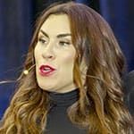 Erin King, with long brown hair, wearing a black top and headset microphone, speaking in front of a dark blurred background.